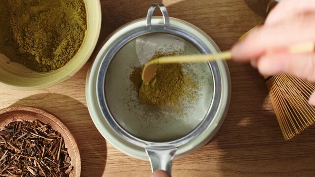 Hand places roasted hojicha powder into mesh sieve and sifts it to refine texture during traditional tea preparation ritual.