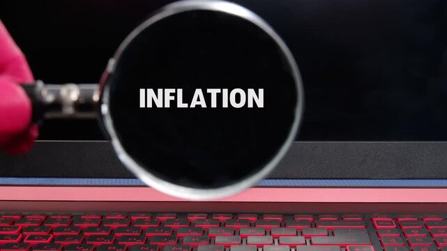 A person uses a magnifying glass to reveal and examine the word inflation on a computer screen. This activity takes place indoors in a study room