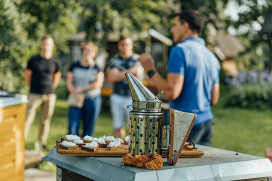 Metal bee smoker and bread samples with white spread sit on a beehive while a small group attends an outdoor beekeeping demonstration in a sunny garden