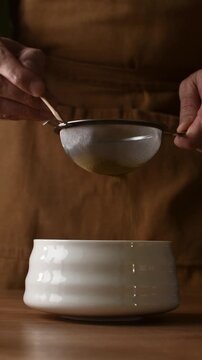 Hands sift roasted hojicha powder through fine mesh sieve into ceramic bowl during traditional Japanese tea preparation.