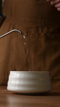 Hot water pours from gooseneck kettle into ceramic bowl to prepare roasted hojicha tea during traditional brewing ritual.