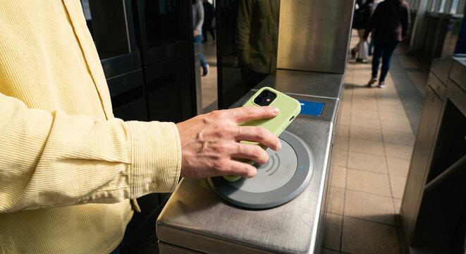 A commuter taps a smartphone on a contactless reader at a transit turnstile, showcasing mobile payment, touchless ticketing, and urban commuting in a busy public transport station