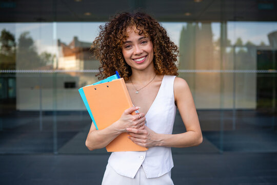 Smiling young corporate woman holding documents outside office