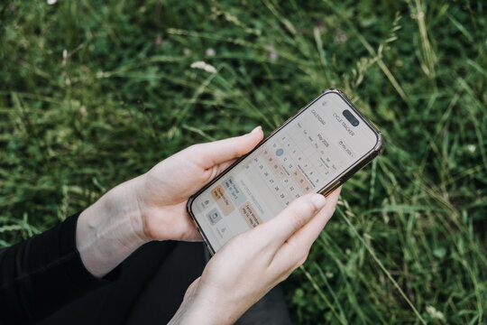 Hands hold smartphone showing calendar interface of cycle tracker application over green grass in park. Femtech calendar interface, menstrual cycle log, ovulation tracking software, 