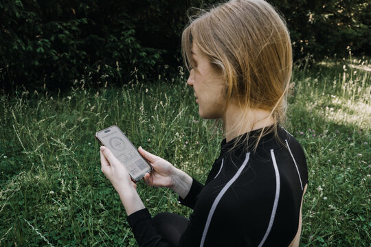 Woman sits in grassy field looking at smartphone screen with health tracking interface. FemTech app tracking, menstrual AI cycle app, women health technology, Hormone tracking app
