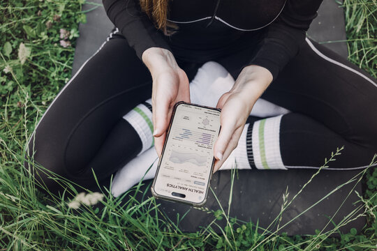 Person sits cross-legged on fitness mat on grass holding smartphone displaying health analytics application. Weekly health summary, wellness data trends, health analytics dashboard, Health report app,