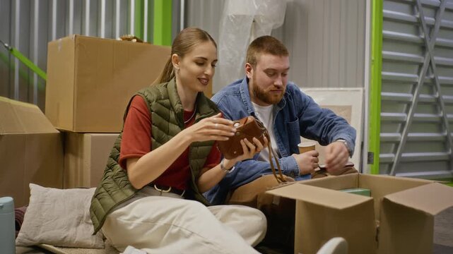 Caucasian young adult couple sitting on floor of self storage unit and unpacking vintage items from cardboard box. Ideal for marketing campaigns promoting relocation or storage services