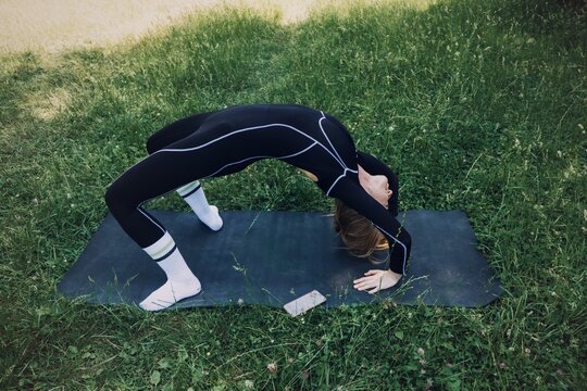 Woman in athletic wear performs wheel pose on mat among grass next to smartphone. Postpartum recovery app, core pelvic strengthening, womens pelvic health technology, Guided pelvic exercises,
