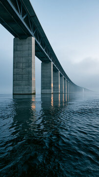 Modern Concrete Bridge Over Water: A long, modern concrete bridge with massive support pillars stretches across calm, rippling water under a hazy, overcast sky.