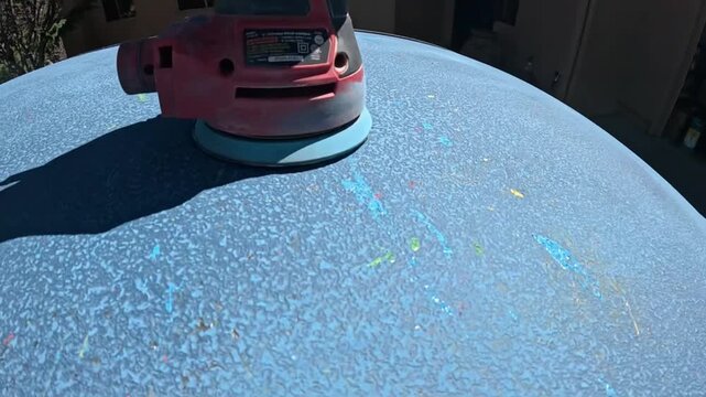 person sanding large blue object outdoors, focused and industrious activity close-up elevated angle.