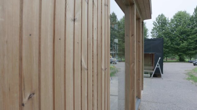 Close view of vertical timber cladding and large glass windows on a small wooden cabin, with reflections, trees, and construction activity in the background