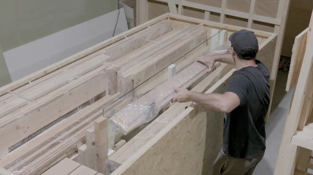 Factory worker packs wrapped wooden construction components into a large shipping crate for export, preparing prefabricated cabin parts for transport