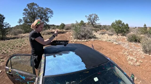 Woman painting car hood in desert beside dark sedan under blue sky with tray and paint splashes near