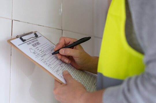 Professional home inspector wearing a safety vest and holding a clipboard with a home inspection report while checking an interior wall for cracks, damage and structural condition