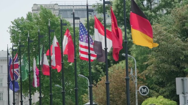 Collection of international flags from various nations waving in the wind against a backdrop of trees and city buildings. Symbolizes global unity and diplomatic relations.
