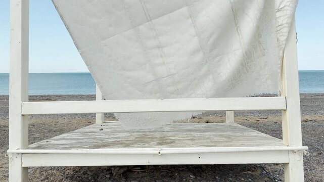 A frayed and cracked white canopy hangs from an empty wooden structure on a deserted beach. The desolate coastal scene stirs a deeply nostalgic and peaceful feeling of time passing.