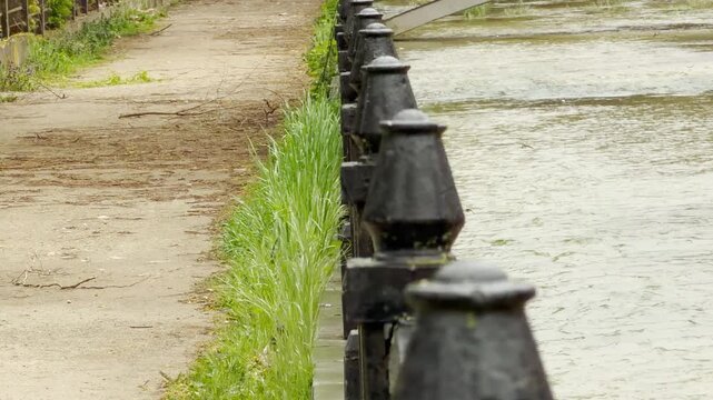 Sturdy iron posts stand guard along a flowing river and dirt walkway. This grey scene feels lonely and quiet under the thick clouds. The damp air creates a somber, peaceful mood.