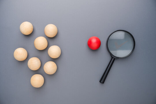 Magnifying glass focusing on a single red wooden ball, representing uniqueness, individuality, talent identification, niche market, outlier detection, recruitment, and business analysis
