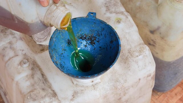 Close-up of an Asian man's hand pouring used cooking oil from a small jerry can into a larger plastic container using a funnel for household waste collection, recycling, and proper disposal.