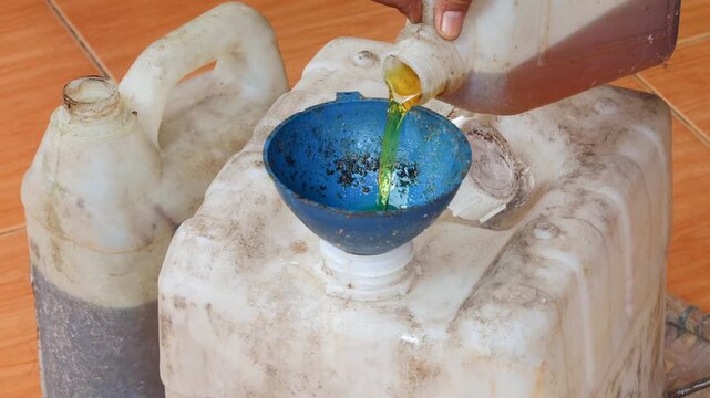 Close-up of an Asian man's hand pouring used cooking oil from a small jerry can into a larger plastic container using a funnel for household waste collection, recycling, and proper disposal.