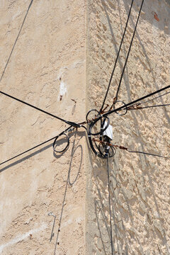 A network of black electrical and telecommunication cables is attached to a rough textured beige stone house corner casting distinct shadows.
