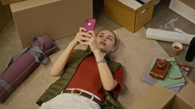 Young adult Caucasian woman lying on floor of storage unit, using smartphone and resting among cardboard boxes. Footage ideal for moving services, self storage promotions, or relocation concepts