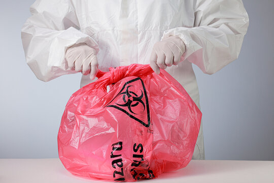Focused worker wearing white protective suit secures red biohazard bag during clinical waste management and disposal process with rubber glove