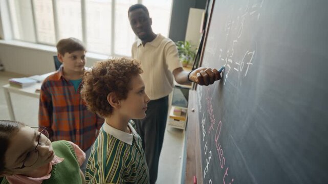 African American male teacher explaining math equations on blackboard to diverse group of children in classroom. Clip is suitable for school advertisements and educational marketing campaigns