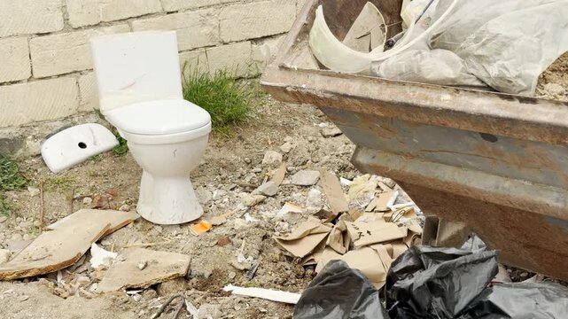 A broken white toilet sits next to messy piles of rubbish and plastic bags. The unpleasant view highlights a dirty, neglected alley filled with scattered construction waste on a bright day.