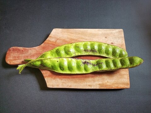 Fresh green petai or stink beans (Parkia speciosa) pods resting on a wooden cutting board against a dark rustic background.