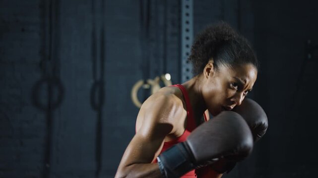 Powerful athletic african american woman with afro hair practicing boxing in a gym. Muscular female boxer throwing punches and training hard in a dark fitness club for self-defense
