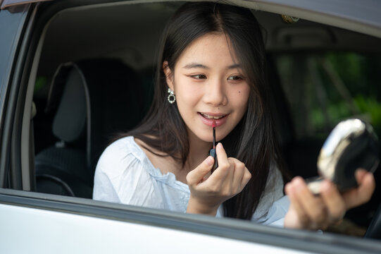 Young Asian woman happily applying lipstick and using a compact mirror while sitting in a parked car