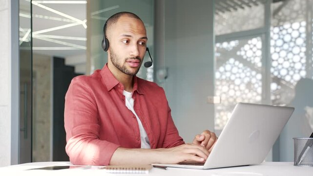 Smiling male corporate operator in headset working in customer support service on helpline telesale talking using laptop in business office. Agent of call center advises client answering questions