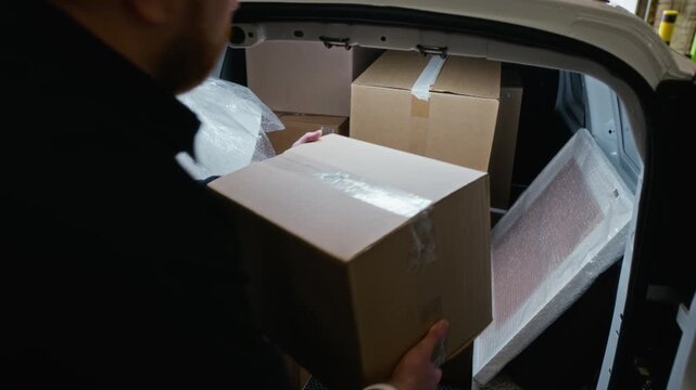 Rear view slowmo of man unloading cardboard box from back of packed delivery van inside storage facility. Clip is ideal for logistics, e-commerce shipping, and moving service advertisements