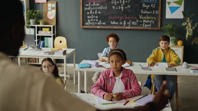 Multiethnic children sitting at desks in classroom raising hands to answer question from African American teacher. Video is ideal for school promotional clips and educational marketing campaigns