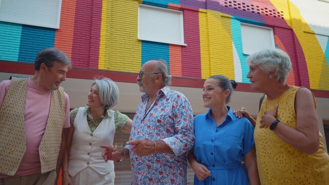 Group of five cheerful senior friends enjoying a sunny day, walking together along a paved path in a modern city park, laughing and talking with one another in a vibrant, friendly atmosphere