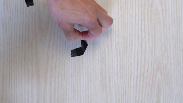Hands removing black tape from wood surface caucasian technician slowly peeling electrical tape marker from pale maple desk closeup fingers lifting sticky adhesive methodical