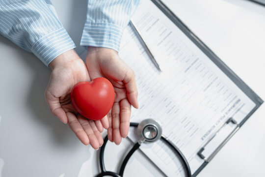 Top view close up doctor hands holding red heart shape model with stethoscope on health insurance policy document desk. Concept of health insurance coverage, cardiology support, and medical care.