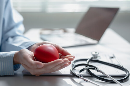 Close up doctor hands holding red heart shape model with stethoscope laptop on insurance document desk. Concept of health insurance coverage, cardiology support, and medical care.