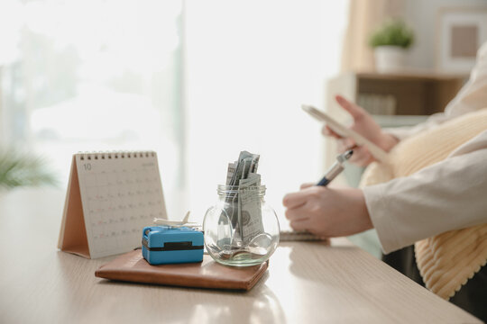 Glass jar filled with money savings, miniature luggage suitcase, toy airplane near hand while writing travel budget. Concept of travel budget savings, vacation financial planning in future.