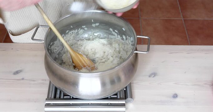 Female hands adding grated Parmigiano Reggiano to hot steaming risotto before mixing with a wooden spoon in a stainless steel pot. Authentic Italian recipe.