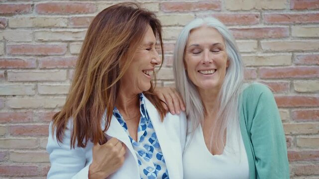 Two happy and stylish senior women friends enjoying their time together. Laughing and smiling heartily while standing against a brick wall. Showcasing a concept of lasting friendship and joy in aging