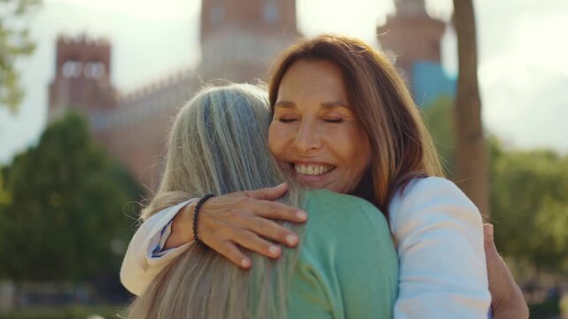Two mature women friends sharing a warm hug outdoors in a sunny park with an old building in the background. The beautiful smiling women enjoy a moment of happiness and genuine friendship