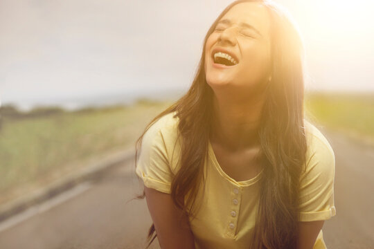young girl laughing with her eyes closed standing on a street with sun in the background