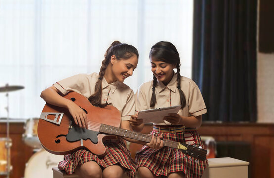 Two happy teenage school girls learning to play guitar looking at musical notes in the music room at school