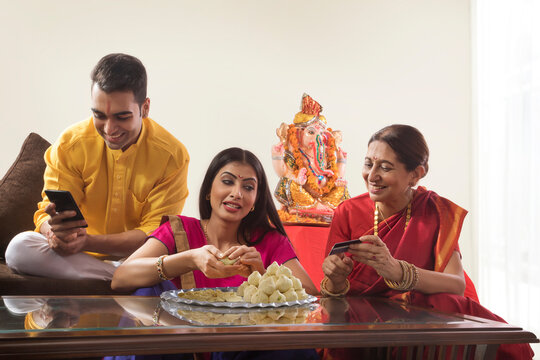 Husband wife sitting together while wife makes modaks and mother in law holds a credit card 