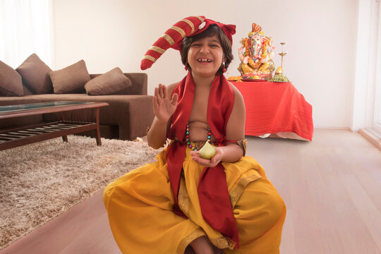 Child sitting dressed up like Ganpati with modak in one hand and Ganpati Idol in background