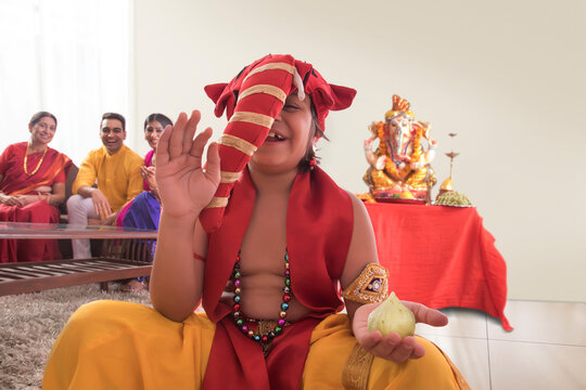 Child sitting dressed up like Ganpati with modak in one hand and Ganpati Idol in background