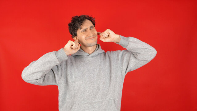 Man in grey hoodie plugging his ears with fingers grimacing while ignoring loud noise on a vibrant red studio background expressing stubborn denial of communication and refusal to listen concept.