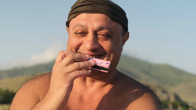Portrait of a cheerful full bodied man with Latin American or Middle Eastern appearance sincerely smiling while playing harmonica shirtless by the sea with mountains. Calm people.